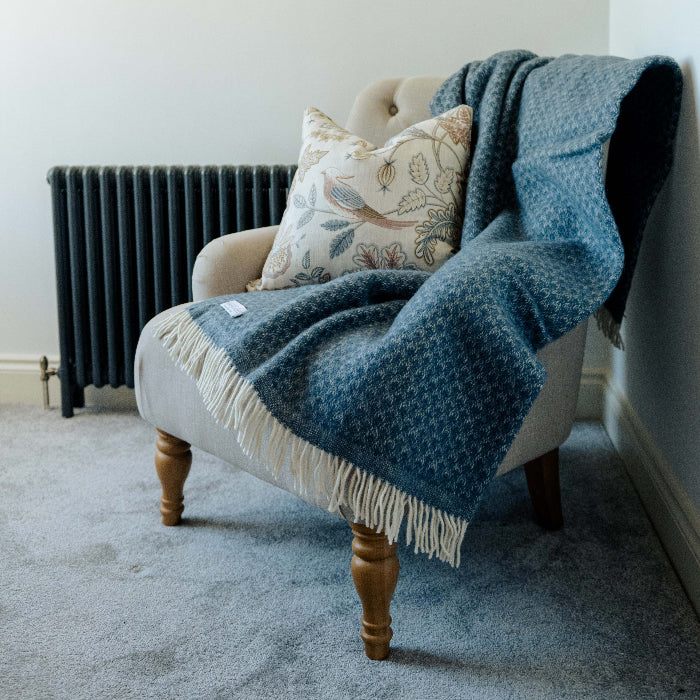 Beige chair with wooden legs and a twilight blue wool throw and patterned pillow against a white wall.