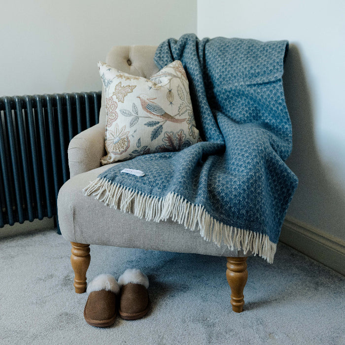 Beige armchair with wooden legs, a twilight blue woollen throw draped on top with a patterned pillow, and slippers on the carpeted floor in front of a radiator.