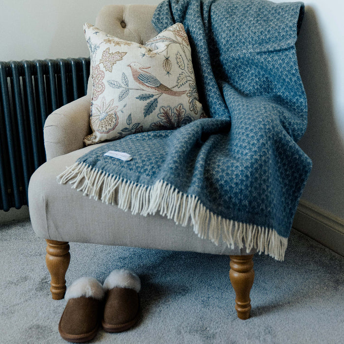 Beige armchair with a twilight blue wool throw, patterned pillow, and brown slippers on a carpeted floor.