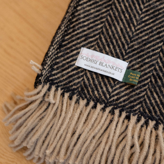 Herringbone patterned blanket in a charcoal and beige colouring with a Solway Blankets label on a wooden surface