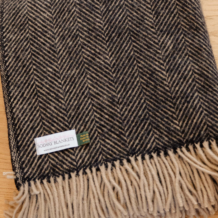 Charcoal and beige herringbone patterned blanket with fringes folded on a wooden surface with a Solway Blankets label