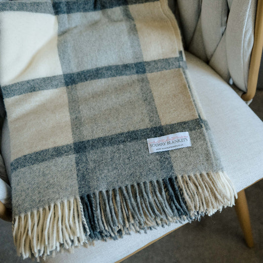 Folded plaid throw with soft grey and white tones draped over a chair with a Solway Blankets label. 